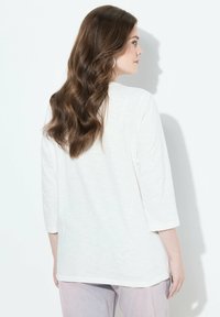 Valkoinen 3/4-hihainen paita, jossa on rento istuvuus, pehmeä tekstuuri ja kevyt kiilto. Näytetty takaa vaaleaa taustaa vasten.