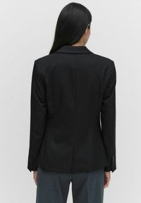 Svart skräddarsydd kavaj med en strukturerad passform, snedskurna lapeller, enkel ventil på baksidan och knappade ärmslut. Slät tyg med minimal detaljer.