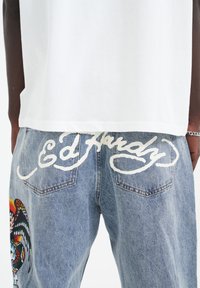 Bílá bavlněná tričko spojená s modrými džínami. Džíny mají bílé vyšívané texty "Ed Hardy" a barevný grafický design na levé noze.