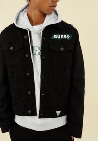 Svart denimjacka med en grön "GUESS" logotyp-patch, knappstängning och två bröstfickor. Buren över en lager-på-lager grå huvtröja.