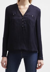 Mörkblå långärmad blus i lätt tyg med V-ringning, två bröstfickor och knappslå, med en avslappnad passform.