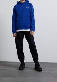 Felpa blu in pile con tasca a marsupio e logo dorato, abbinata a pantaloni della tuta neri e sneakers nere su una superficie grigia.