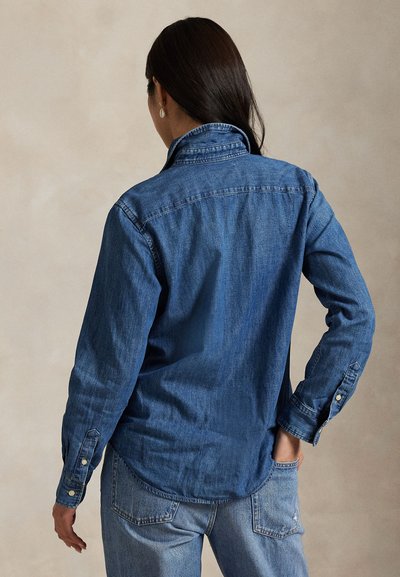 Femeie cu păr lung și închis la culoare, purtând o cămașă din denim albastru și blugi de un albastru deschis, stând cu o mână în buzunarul din spate.