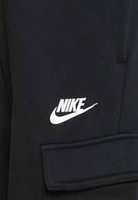 Černé tepláky s texturovaným povrchem, s bílým logem Nike a náprsní kapsou na boku.