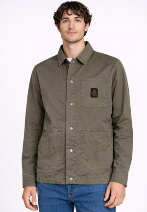 Młody mężczyzna w oliwkowo-zielonej kurtce zapinanej na guziki z naszywką na kieszeni piersiowej oraz w niebieskich dżinsach, stoi na jednolitym jasnym tle.