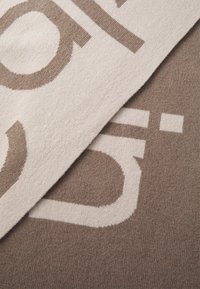 Coperta lavorata a maglia con testo intrecciato in tonalità crema e marrone. Texture liscia con un motivo sottile e blocchi di colore a contrasto.
