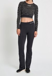 Kvinna i en figursydd, långärmad, zebra-mönstrad cropped topp och mörka träningsbyxor, stående mot en neutral bakgrund, iförd svarta sneakers.