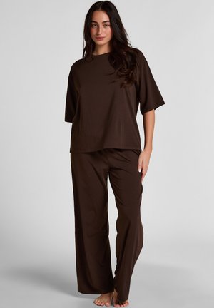 Frau steht barfuß und trägt ein locker sitzendes, dunkelbraunes Kurzarm-Top und passende weit geschnittene Hosen vor hellgrauem Hintergrund.