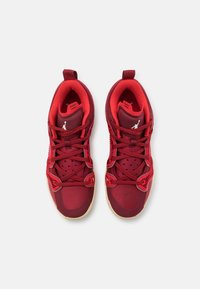 Paire de baskets rouges avec des lacets à motifs et un logo blanc sur des languettes rembourrées, vues de dessus sur un fond clair uni.
