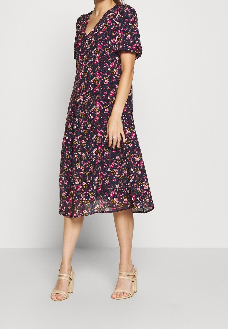Květinové midi šaty s tmavě modrým pozadím, krátkými nabíranými rukávy, výstřihem do V a splývavým lemem, zdobené růžovými a žlutými květinovými vzory.