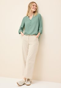 Blusa verde chiaro a maniche lunghe con scollatura a V e dettagli con bottoni, abbinata a pantaloni beige a gamba larga e sneakers grigie. Tessuto dalla consistenza liscia.
