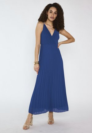 Donna con capelli ricci che indossa un abito blu lungo plissettato senza maniche e sandali dorati, posando con una mano sul fianco su uno sfondo semplice.