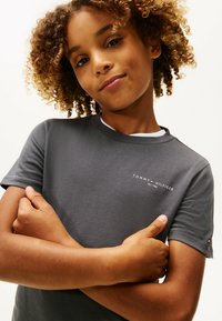 Barn med krøllet hår iført en mørkegrå Tommy Hilfiger t-shirt, armene krydset, ser selvsikkert ind i kameraet mod en ensfarvet baggrund.