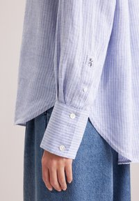 Camicia a righe blu chiaro con maniche lunghe e polsini abbottonati in tessuto morbido. Presenta un piccolo dettaglio con logo vicino al polsino. Indossata sopra una gonna di jeans.
