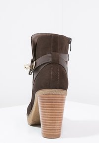 Brun mocka ankelstövel med en kurvad, staplad klack. Har en läderrem med en guldtonad spänne som dekor. Sidodragkedja.