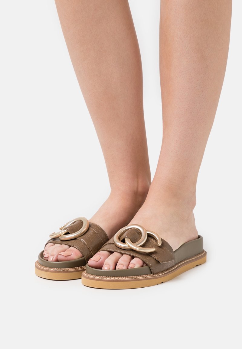 Sandales marron à enfiler avec deux accents en boucle métallique, dessus en cuir texturé et semelle plate avec une base en caoutchouc beige.