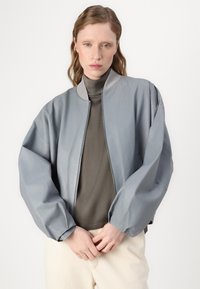 Casaco bomber cinza claro com textura suave, colarinho canelado e mangas volumosas. Usado sobre uma blusa de gola alta cinza escuro.