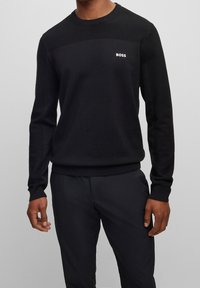 Svart stickad tröja med texturerad ok och ribbade ärmslut. Litet vitt logo på bröstet. Kombinerad med svarta byxor.