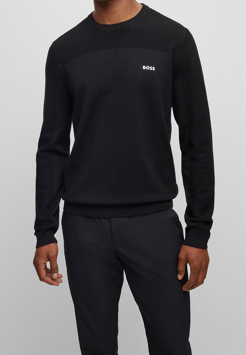 Svart stickad tröja med texturerad ok och ribbade ärmslut. Litet vitt logo på bröstet. Kombinerad med svarta byxor.