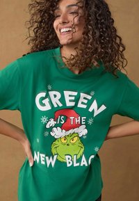 Groene katoenen T-shirt met een Grinch-illustratie en een rode Santa-hat. Tekst luidt "GREEN IS THE NEW BLACK" in vetgedrukte witte letters.