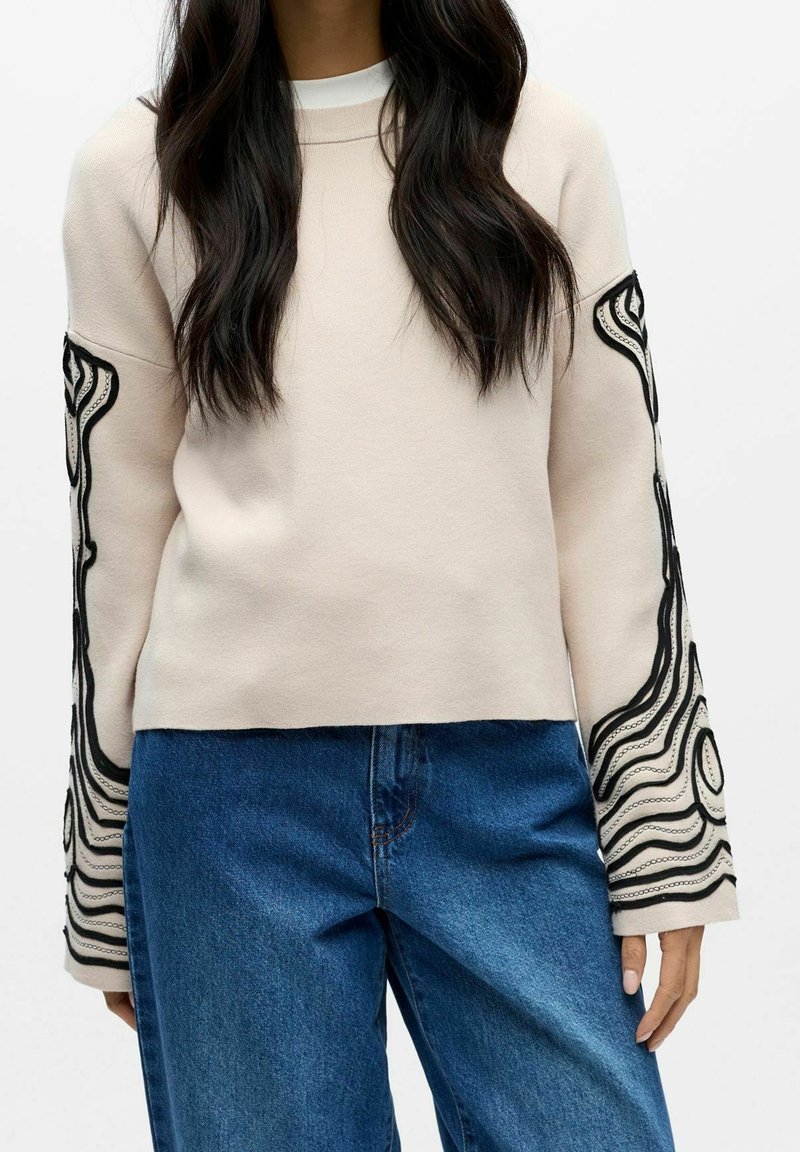 Femme portant un haut beige à manches longues avec des motifs tourbillonnants noirs sur les manches et un jean en denim bleu, debout devant un fond uni.