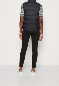 Person, der eine schwarze Steppweste, schwarze enge Jeans, ein schwarz-weiß gestreiftes Kurzarmshirt und weiße Sneaker trägt, steht gegen eine weiße Wand.