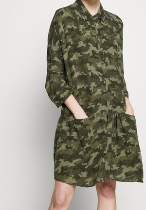 Mujer que lleva un vestido verde de camuflaje con botones, de corte holgado hasta la rodilla, con las mangas remangadas y las manos en los bolsillos delanteros.