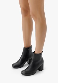 Botine negre din piele cu un finisaj neted, vârfuri rotunde și un toc pătrat de 10 cm; au un fermoar pe lateral pentru a fi ușor de purtat.