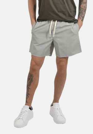 Mężczyzna w szarych, wzorzystych szortach ze sznurkiem, oliwkowo-zielonej koszuli, białych trampkach i tatuażami na ramionach oraz lewej nodze, stoi z rękami w kieszeniach.