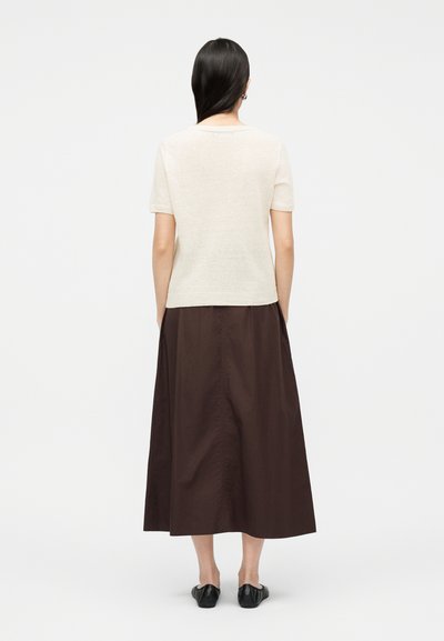 Femme aux longs cheveux foncés portant un haut crème à manches courtes, une jupe midi marron foncé et des chaussures plates noires, debout de dos sur un fond blanc.