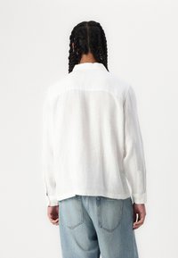 White linen shirt with a relaxed fit, buttoned cuffs, and a back yoke detail. Paired with light blue denim jeans.