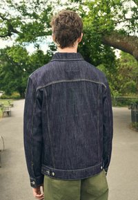 Mørk denimjakke med lodrette striber, der har kravehals, trykknaplukninger og sidelommer. Båret over olivengrønne bukser.