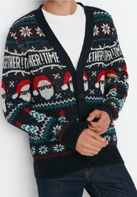 Uomo che indossa un cardigan scuro con bottoni e motivi natalizi come facce di Babbo Natale, fiocchi di neve, stelle e il testo "INSIEME! TEMPO" su una maglietta bianca e jeans.
