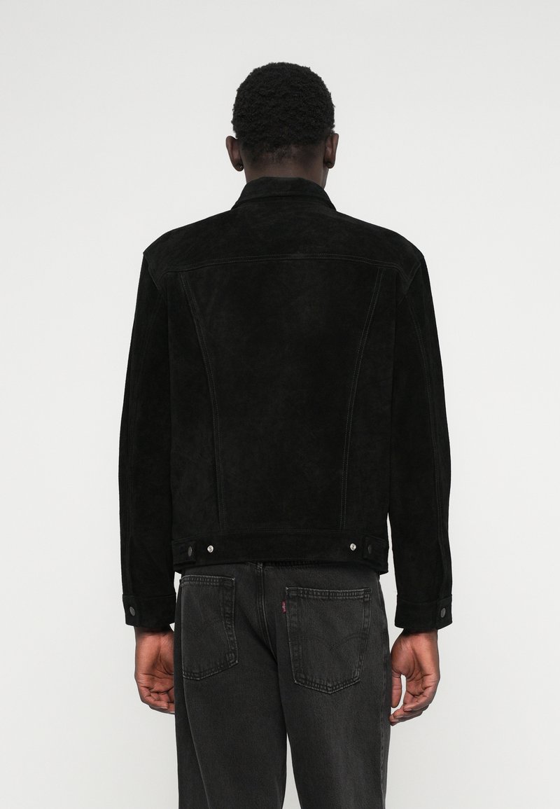 Giacca in suede nera con colletto classico, spalla posteriore e bottoni a pressione, abbinata a jeans scuri. Presenta una finitura testurizzata e una silhouette strutturata.