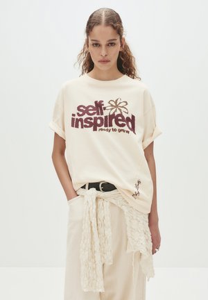 Cream T-shirt with burgundy embroidered text "self inspired" and a flower. Loose fit, round neckline, rolled sleeves, paired with light pants.
