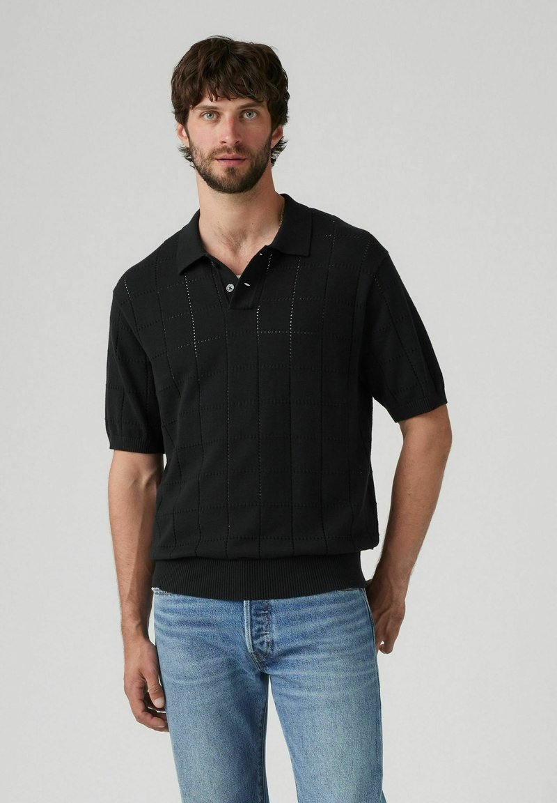 Camisa polo negra de manga corta con un patrón de cuadrícula texturizado, que cuenta con un cuello y una tapeta de botones. Llevada con jeans de mezclilla azules.
