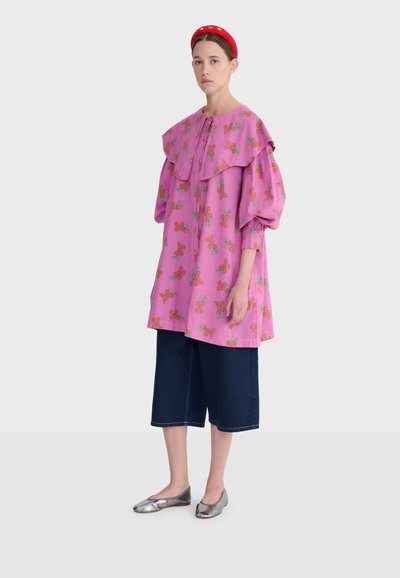 Femme portant un grand chemisier rose à fleurs avec un col large, un pantalon large bleu marine raccourci, un serre-tête rouge et des ballerines argentées, debout devant un fond blanc.