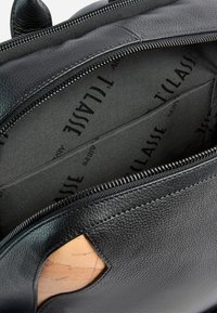 Interno di una borsa in pelle nera con chiusura a zip, caratterizzato da una superficie testurizzata e una fodera decorata con il marchio 'TILALISE' e una tasca.