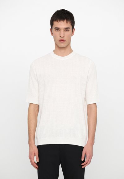 Hellfarbener, gerippter Strickpullover mit kurzen Ärmeln und Rundhalsausschnitt, kombiniert mit dunklen Hosen. Die Textur ist weich und leicht durchsichtig.