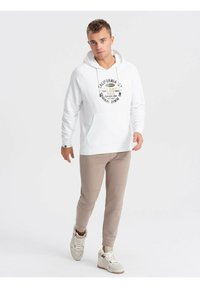 Sudadera blanca con bolsillo frontal y estampado gráfico, combinada con joggers beige ajustados y zapatillas blancas. Tejido suave, diseño casual.