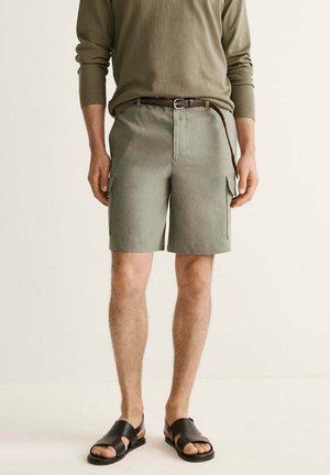 Mann trägt olivgrühes langärmliges Hemd, passende Cargoshorts mit Seitentaschen, braunen geflochtenen Gürtel und schwarze Sandalen mit offenen Zehen, steht drinnen.