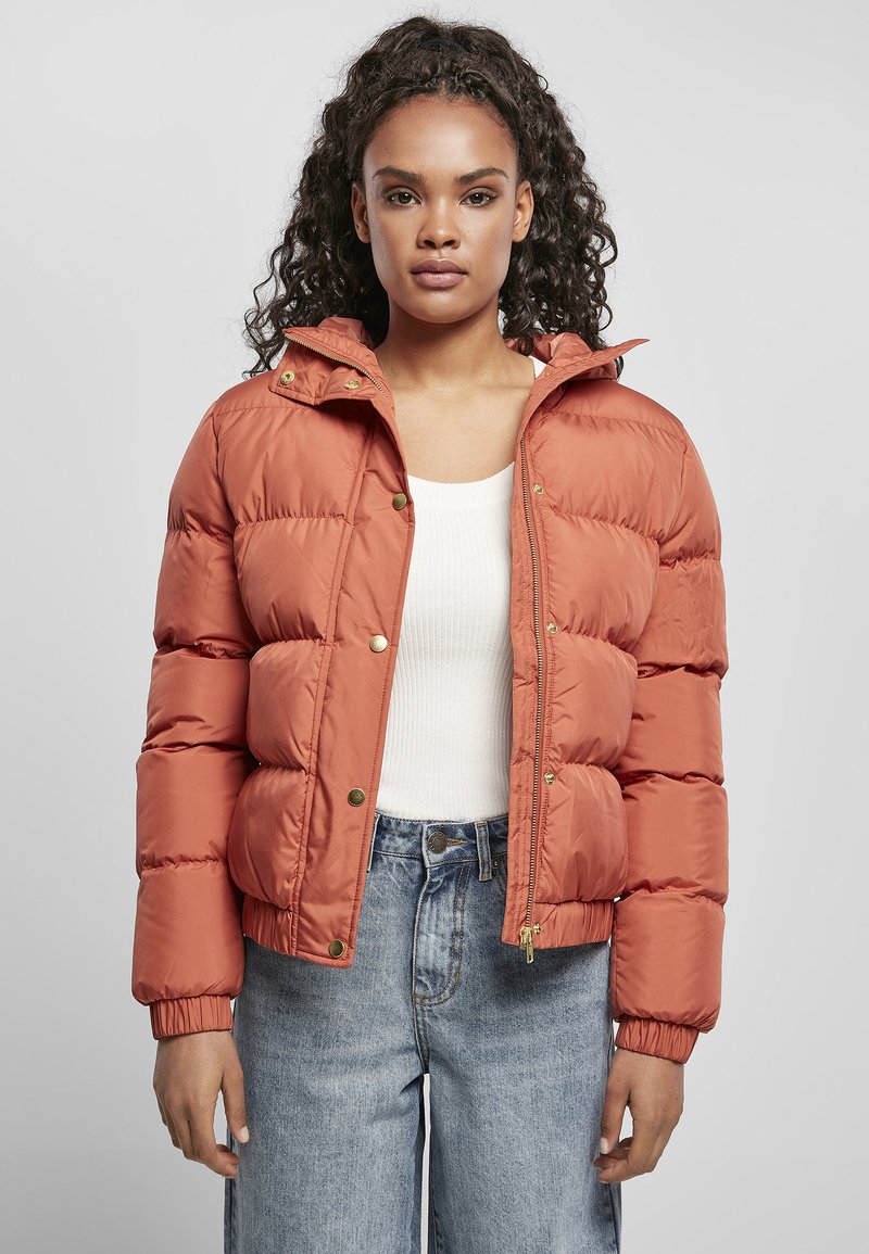 Ulllete, oransje jakke med glidelås foran og trykknapper, med hette og elastiske mansjetter, kombinert med en hvit topp og jeans.