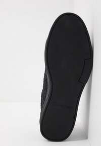 Černá gumová podrážka boty s texturovaným vzorem a vyraženým názvem značky "Calvin Klein." Obsahuje hladký okraj a různorodý dezén.