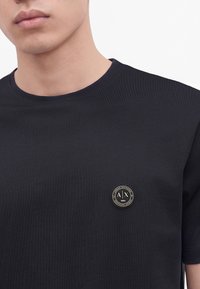 Gros plan d'une personne portant un t-shirt noir avec un patch rond du logo Armani Exchange sur la poitrine.