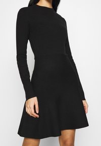 Svart långärmad klänning med fitted överdel och utsvängd kjol. Tillverkad av stickad tyg, har en rundad halsringning och en jämn struktur.