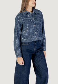 Giacca di jeans cropped di colore blu scuro, adornata con paillettes luccicanti, dotata di colletto, chiusura con bottoni e maniche lunghe. Abbinata a jeans a gamba larga.