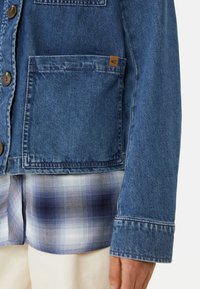 Jeansowa kurtka w niebieskim odcieniu, z kieszonką na piersi, brązowymi guzikami oraz koszulą w kratę pod spód w odcieniach niebieskiego i białego.