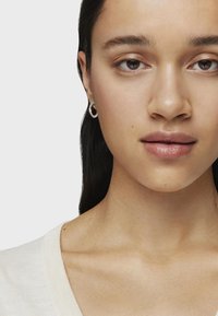Pendiente de aro plateado llevado en la oreja izquierda. Diseño circular y suave; el modelo tiene un tono de piel medio y cabello oscuro, y lleva un suéter claro.