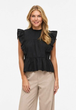 Blondynka uśmiechająca się, ubrana w czarną, falbaniaste, bezrękawnikową bluzkę i beżowe spodnie, stojąca na tle białego tła.