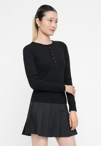 Jonge vrouw met lang haar in een zwart geknoopte langmouwen top en een donkere geplooide rok, staand tegen een witte achtergrond.